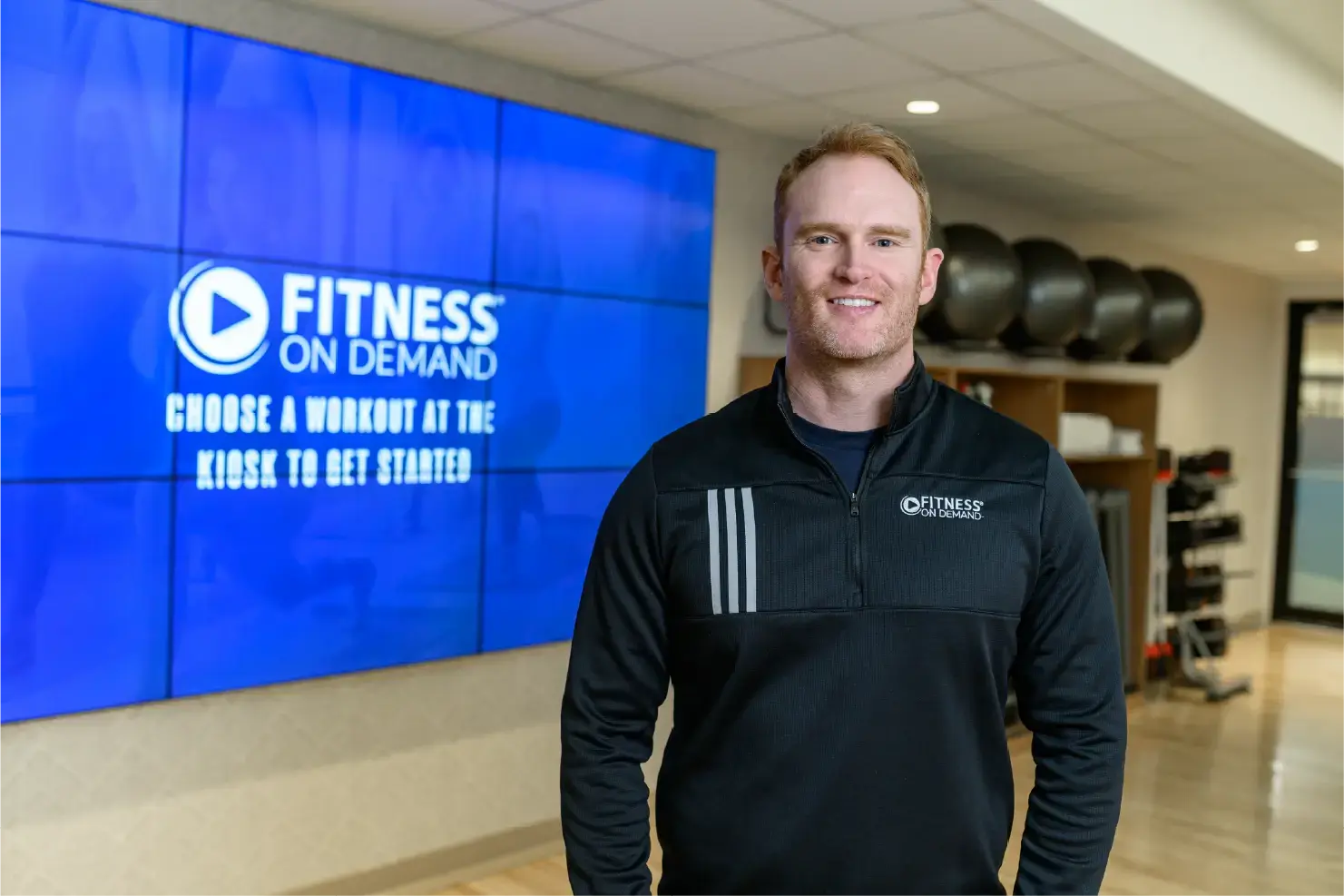 Andy Peat, CEO, in a black Fitness On Demand jacket stands smiling in a gym, with exercise balls and a large screen displaying Fitness On Demand in the background.