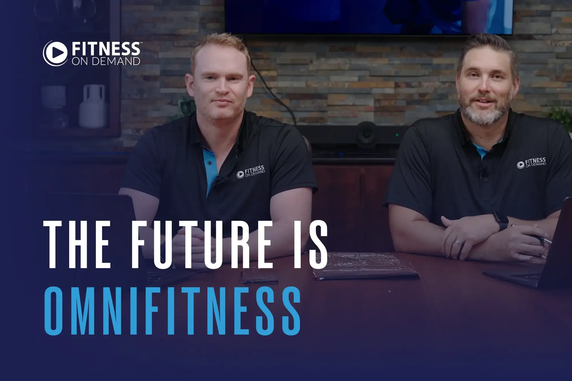 Andy Peat and Todd Wiginton wearing matching Fitness On Demand shirts sit at a desk with laptops and papers. Text overlay reads "The Future is Omnifitness".