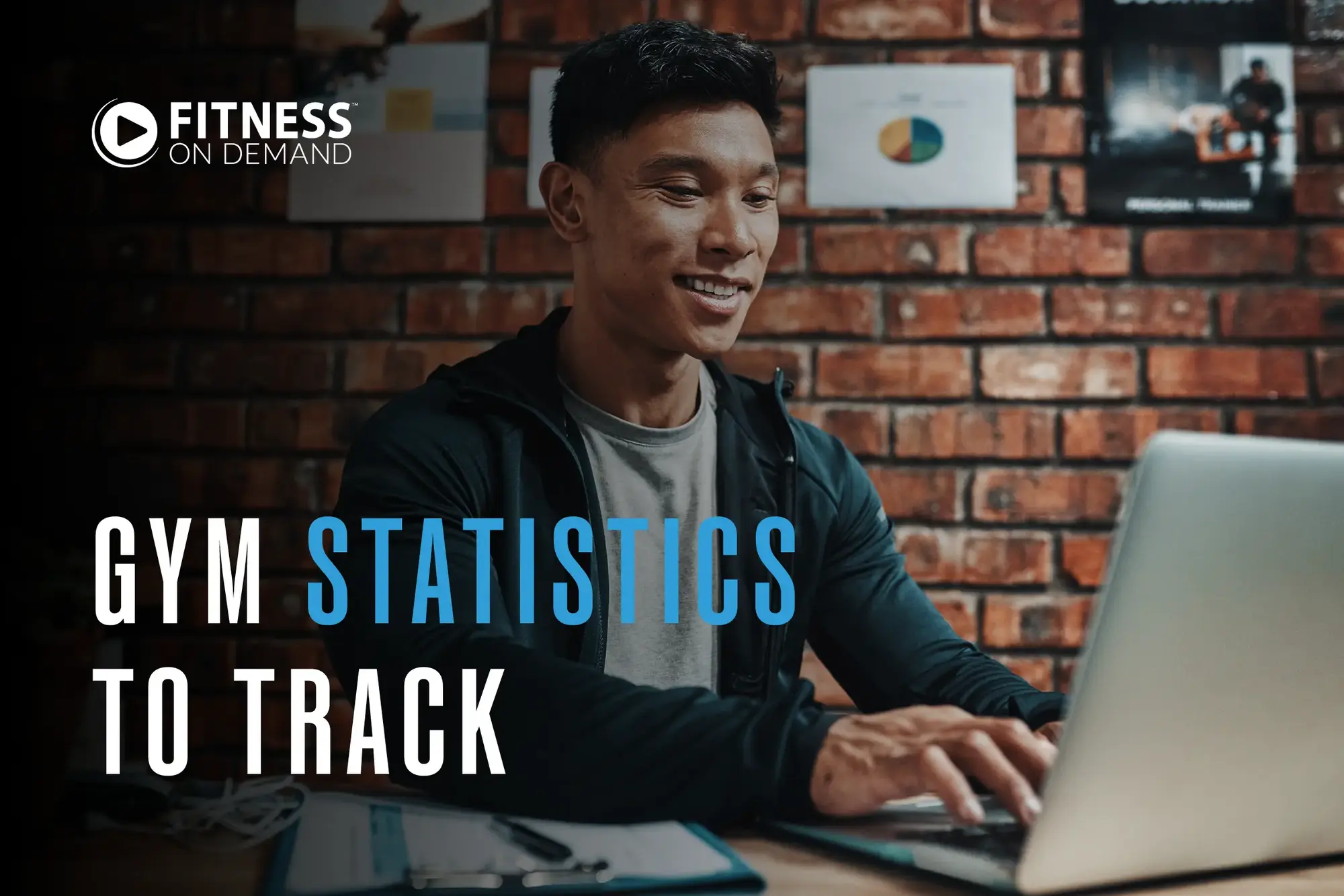 A man sits at a desk smiling and typing on a laptop, with charts on a brick wall behind him. Text on the image reads "Gym Statistics to Track"