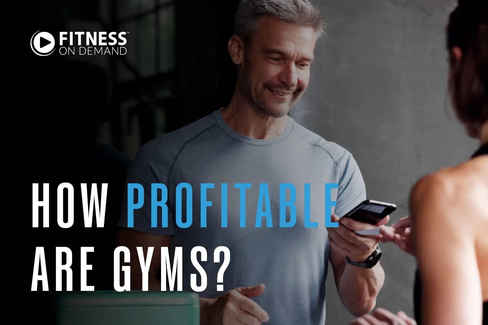 Smiling man in workout clothes holds a smartphone while speaking to another person. Text reads: How profitable are gyms? Fitness On Demand