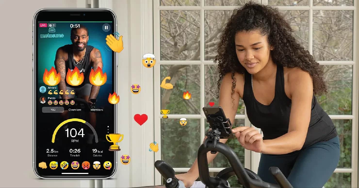 A woman rides an exercise bike indoors, looking at her phone. An app on the phone displays a virtual cycling instructor, workout stats, and animated reactions like flames, trophies, and emojis.