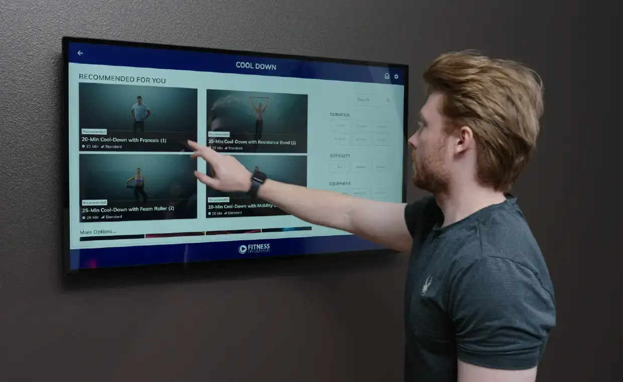 A man touches a wall-mounted touchscreen displaying cool down workout videos, selecting one from recommended options.