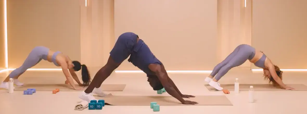 Three people practice yoga indoors, performing the downward dog pose on mats. Dumbbells and water bottles are nearby, and the room has soft lighting and a minimalist design.