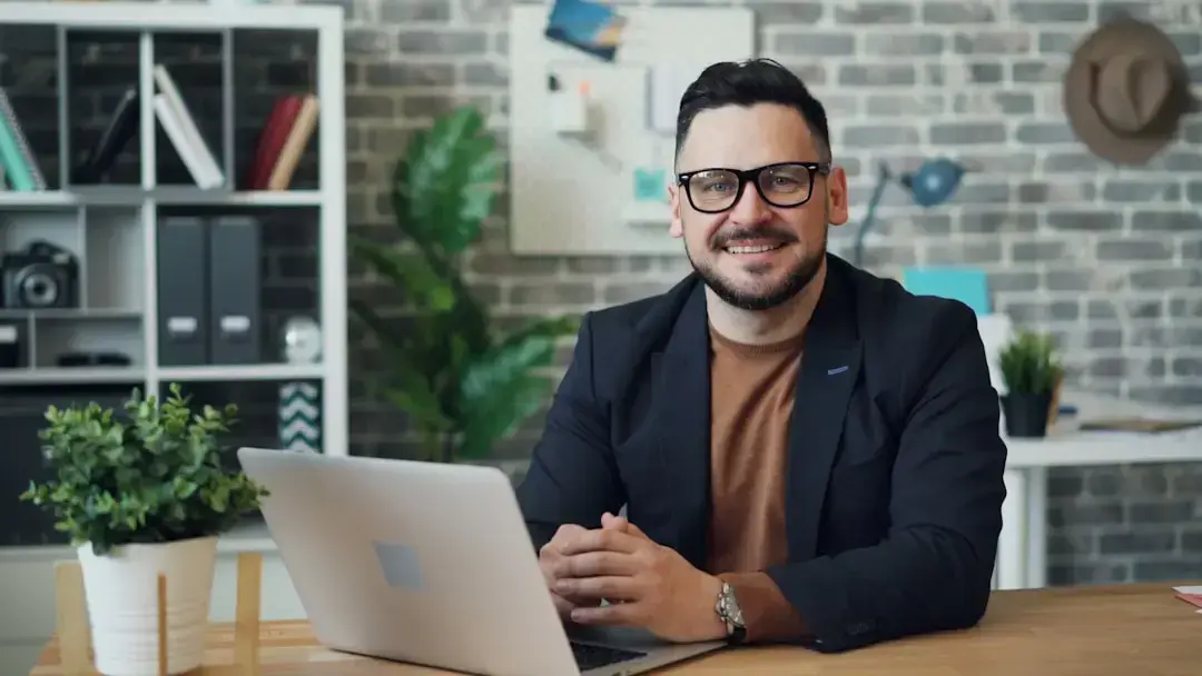 A man wearing glasses and a blazer sits at a desk with a laptop, smiling at the camera. Behind him are shelves, a plant, and a corkboard on a brick wall.