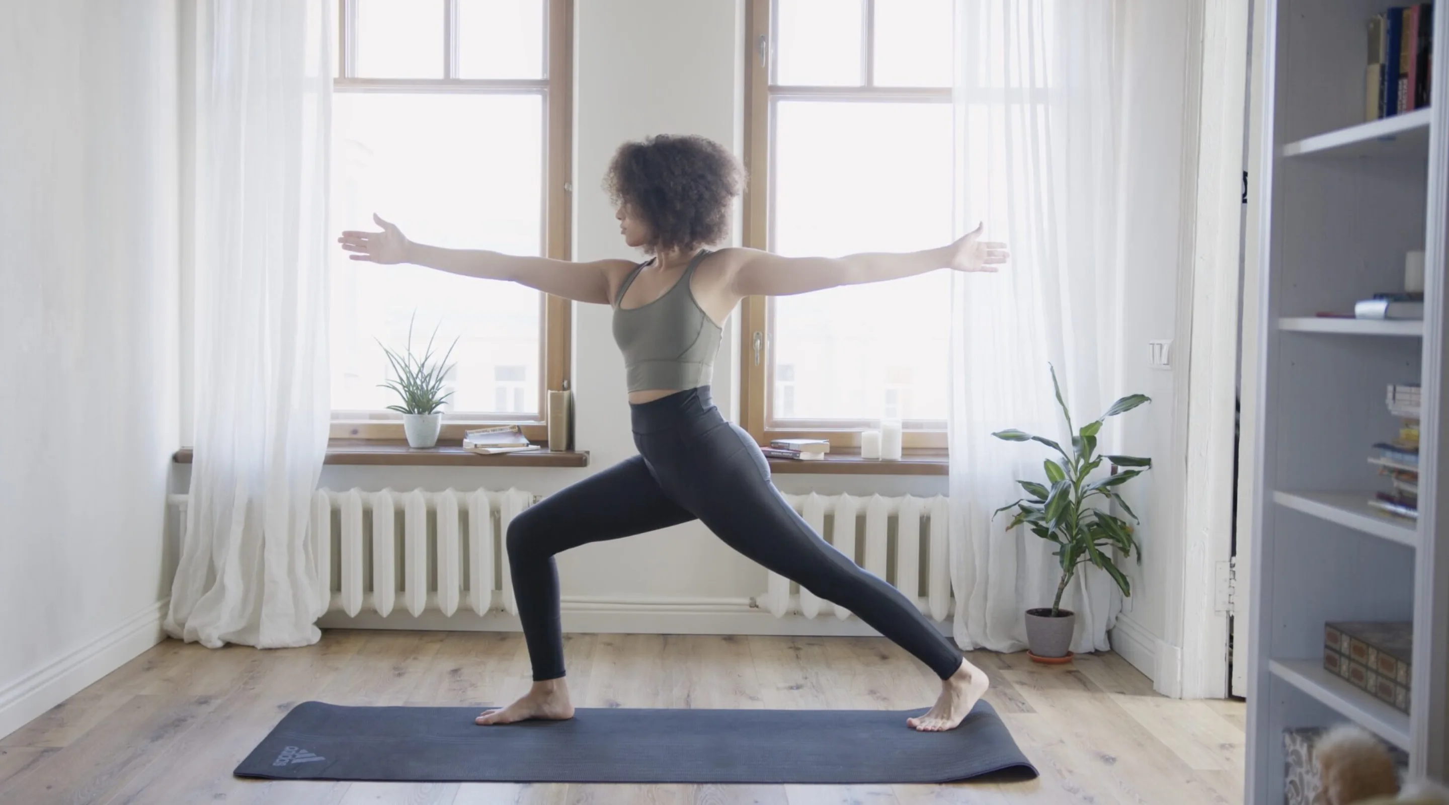 A person practices yoga indoors, standing on a mat in Warrior II pose with arms extended. Sunlight streams through two large windows behind them, and there are potted plants in the room.