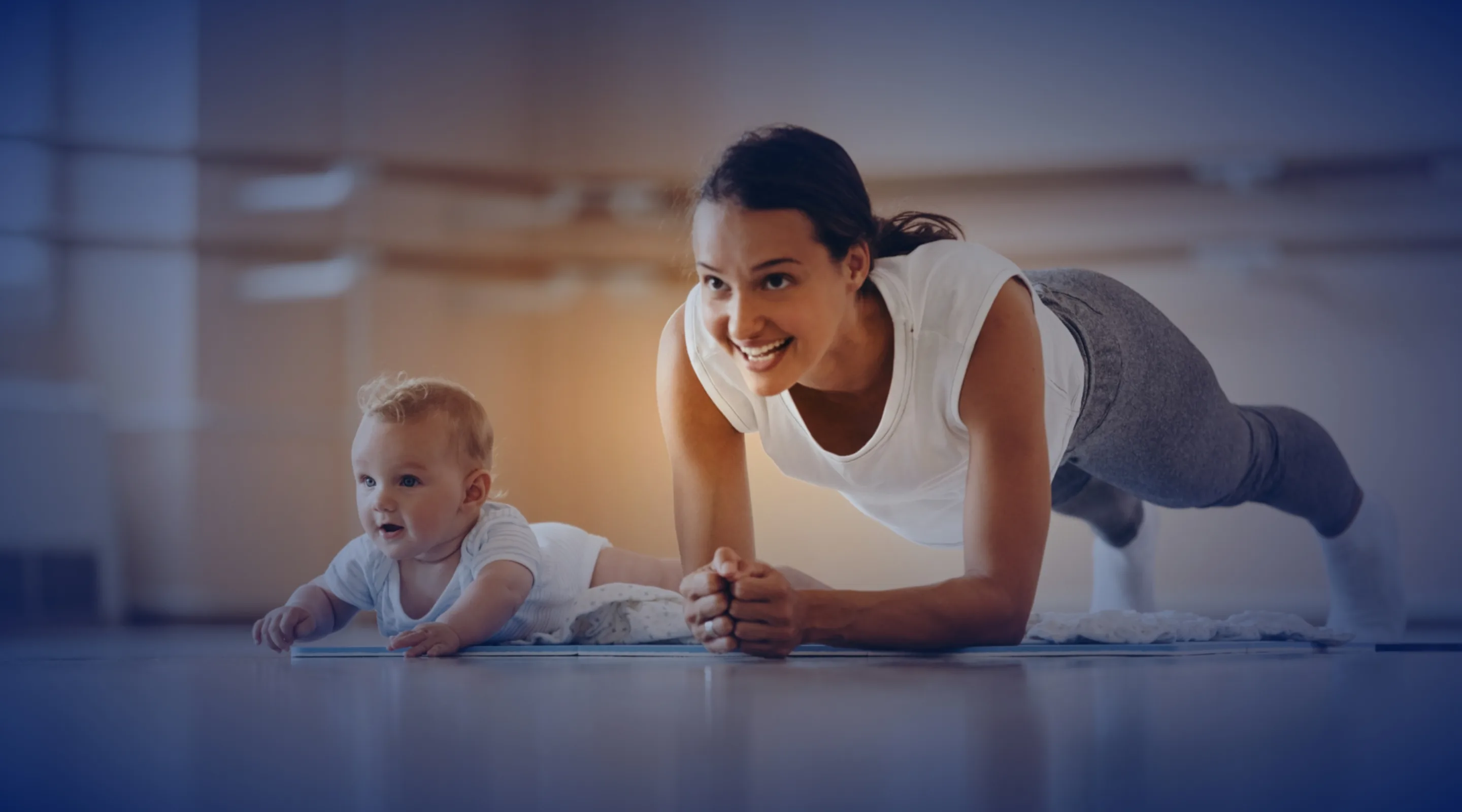 A smiling woman in workout clothes does a plank on a yoga mat beside a baby lying on their stomach in a bright, spacious room.