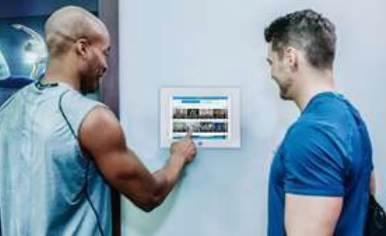 Two men in athletic wear stand by a wall-mounted touchscreen, with one man pointing at the screen and both smiling, possibly selecting a workout or gym activity.