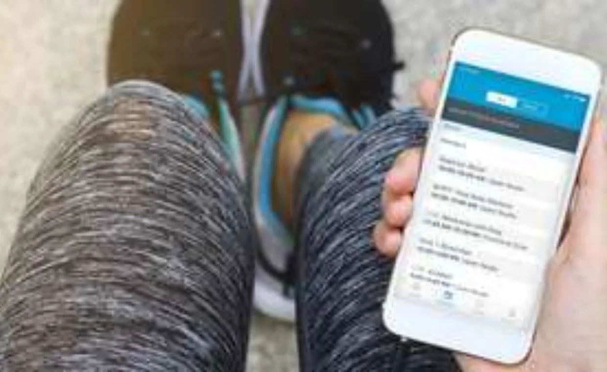 A person in athletic wear holds a smartphone displaying a fitness or health app while seated, with black running shoes visible in the background.