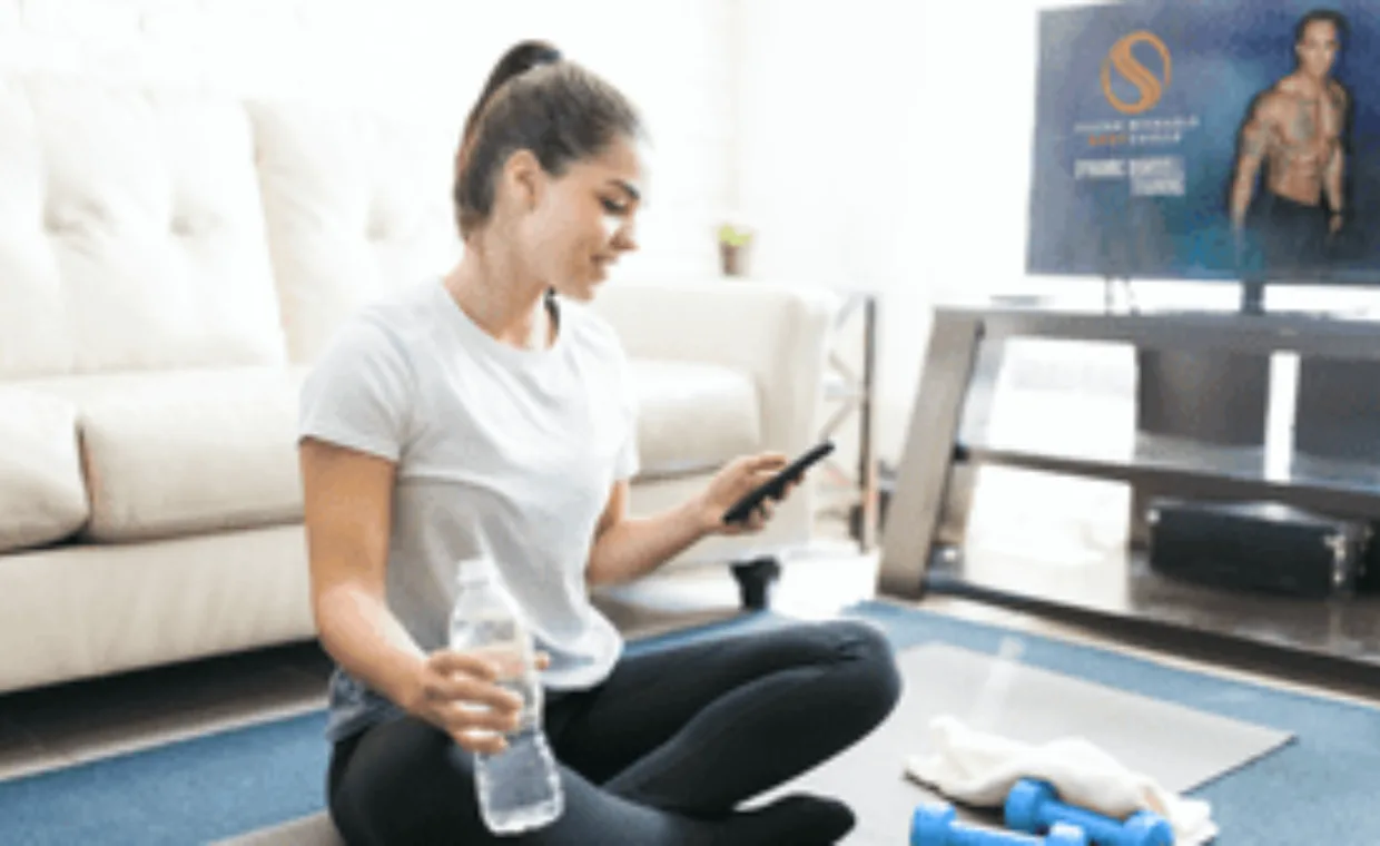A woman in workout clothes sits on a mat in her living room, holding a water bottle and looking at her phone. Dumbbells and a towel are nearby, and a fitness program is displayed on the TV.