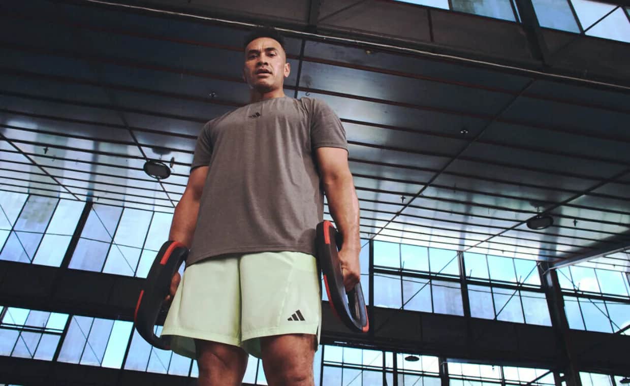 A man in a gym setting stands holding two weight plates by his sides, wearing a gray T-shirt and light green shorts, with large windows and a high ceiling in the background.