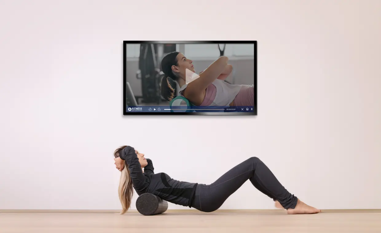 A woman lying on a foam roller in a minimal room follows an exercise video displayed on a wall-mounted TV, mimicking the workout shown on the screen.